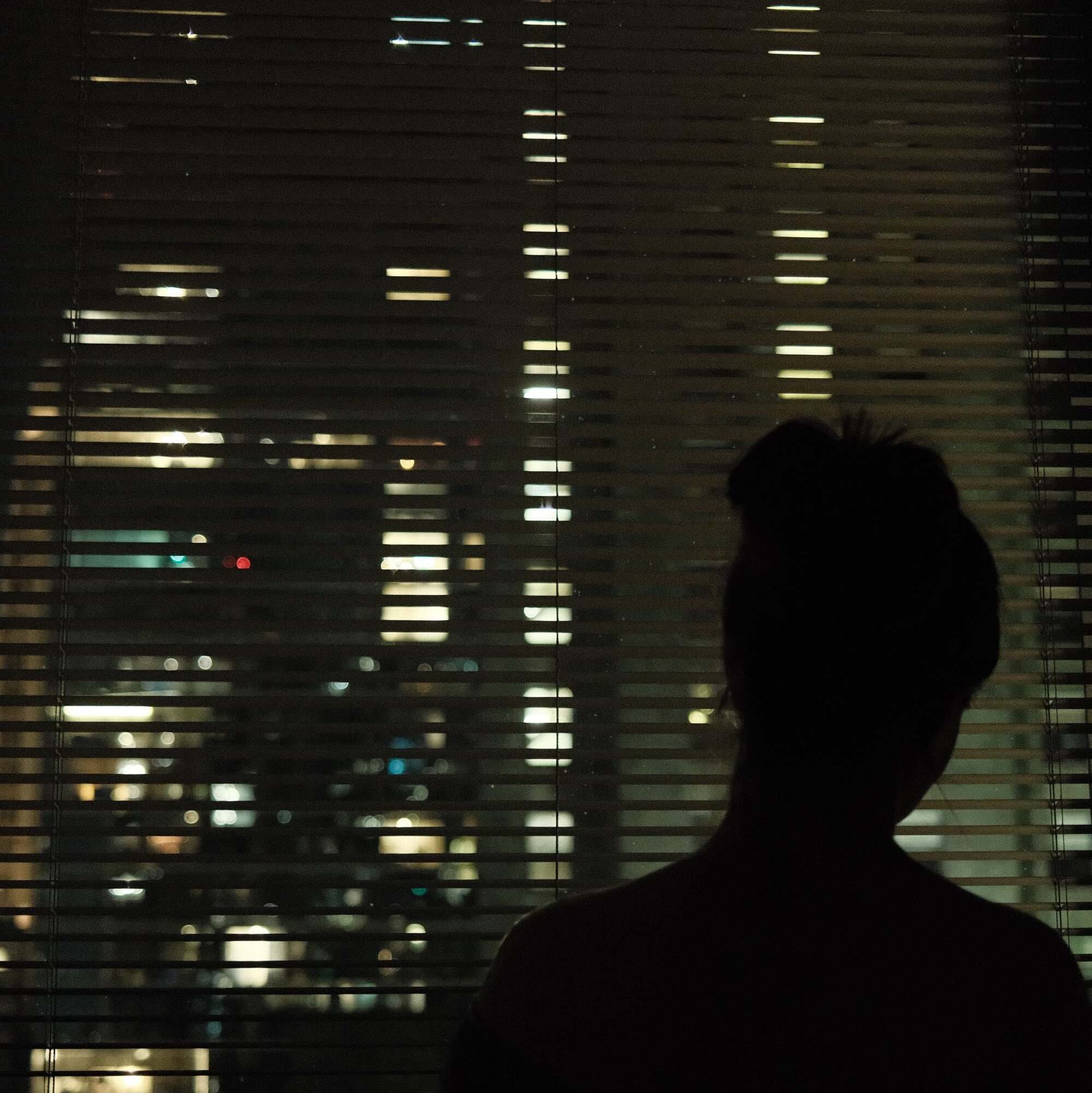 Silhouetted figure watching city lights through venetian blinds at night
